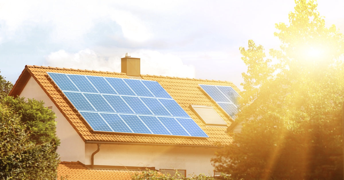 Several large solar panels are installed on the tiled roof of a residential home. The sun shines brightly on the panels.