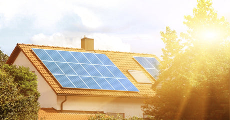 Several large solar panels are installed on the tiled roof of a residential home. The sun shines brightly on the panels.
